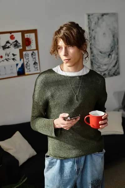 A young man stands at home, holding a coffee cup and looking thoughtfully at his phone. — Stock Photo