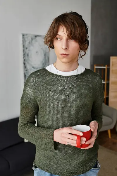 En un ambiente acogedor en casa, un joven se relaja con una taza caliente, profunda en el pensamiento mientras estudia. - foto de stock