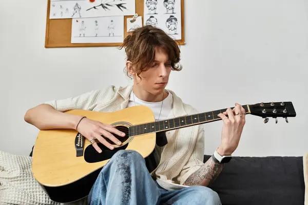 Giovane uomo si siede su un divano, strumming una chitarra pensieroso in un'atmosfera accogliente casa. — Foto stock