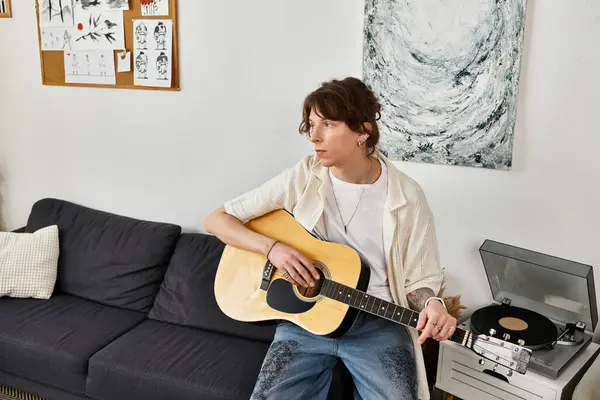 Un giovane uomo si siede sul divano, suonando la chitarra pensieroso mentre si gode il suo ambiente domestico. — Foto stock