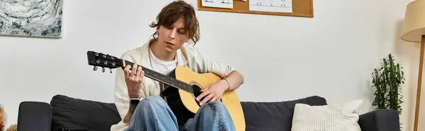 A young man sits comfortably on a sofa at home, strumming a guitar and focusing on music. — Stock Photo