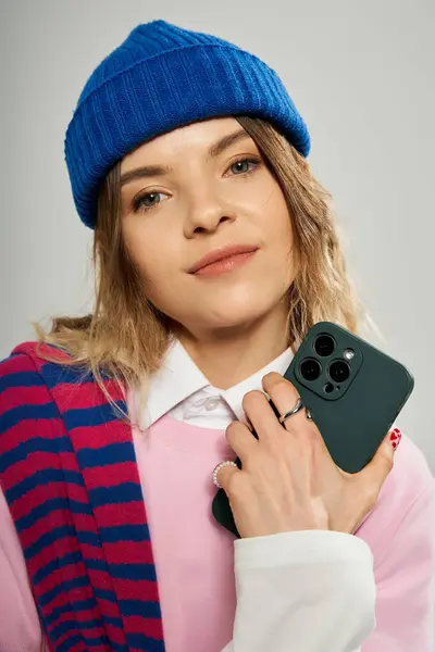 A pretty young woman wearing a blue beanie holds her phone, radiating confidence in a studio. — Stock Photo