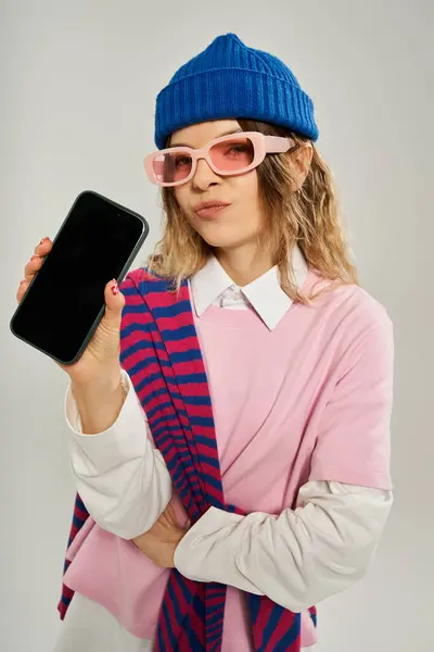 A young woman models her stylish outfit with a blue beanie and fun sunglasses indoors. — Stock Photo