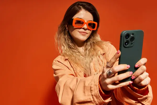 A young woman poses confidently in a bold outfit against an orange backdrop while taking selfies. — Stock Photo