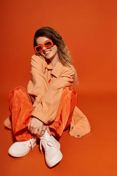 A stylish young woman sits playfully on an orange backdrop, capturing a cheerful vibe. — Stock Photo