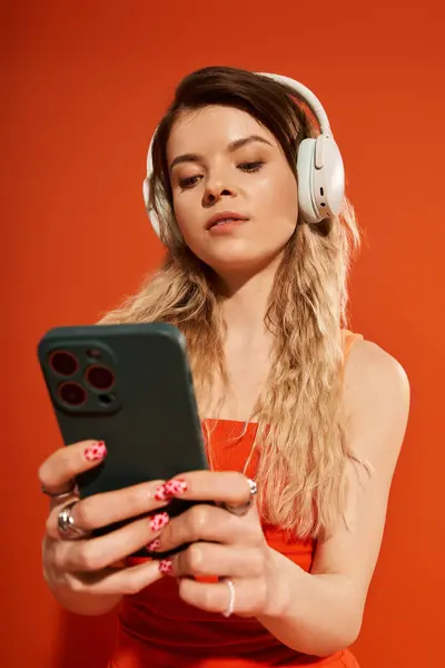 A pretty young woman is listening to music while scrolling on her phone against an orange backdrop. — Stock Photo