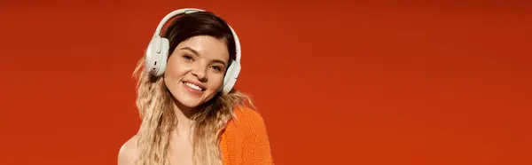 A stylish young woman joyfully models a trendy outfit and headphones against an orange background, banner — Stock Photo