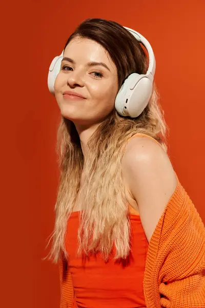 Eine stylische junge Frau lächelt mit Kopfhörern vor leuchtend orangefarbener Kulisse. — Stockfoto