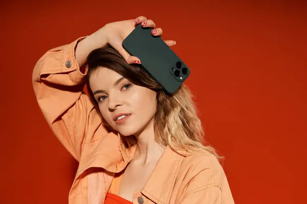 Una joven confiada muestra su elegante atuendo y teléfono sobre un fondo naranja. - foto de stock