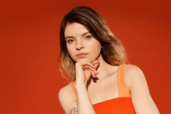 Una mujer joven con estilo posa con confianza con una expresión reflexiva sobre un fondo naranja. - foto de stock
