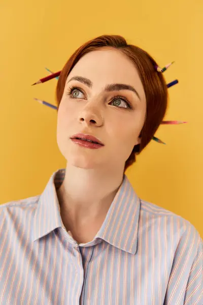 Uma jovem mulher olha para cima, lápis coloridos em seu cabelo, contra uma parede amarela brilhante. — Fotografia de Stock