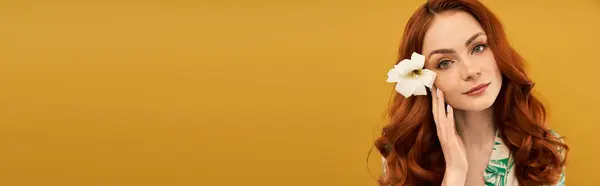 A lovely young woman touches her face gently while showcasing a beautiful flower in her hair. — Stock Photo