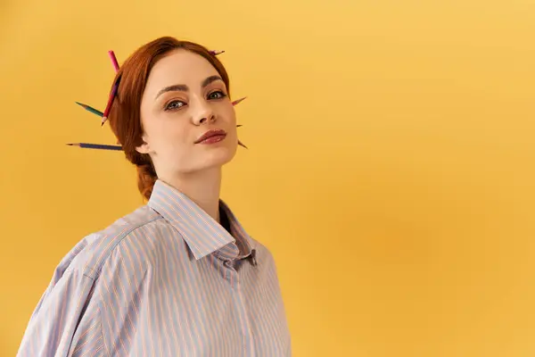 Una joven posa confiadamente sobre un fondo amarillo brillante con lápices en el pelo. - foto de stock