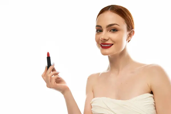 Una hermosa joven sonríe con confianza mientras sostiene un vibrante lápiz labial rojo en un entorno brillante. - foto de stock