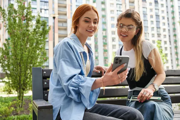 Two stylish young women sit on a bench outdoors, enjoying each others company and conversation. — Stock Photo