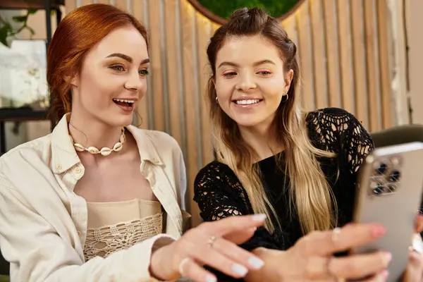 Due amici sorridono e condividono un momento insieme utilizzando uno smartphone in un caffè alla moda. — Foto stock