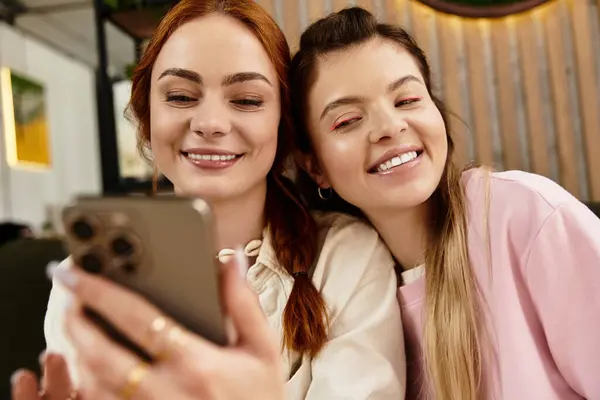 Dos mujeres jóvenes comparten risas mientras capturan un momento alegre en el teléfono en un café moderno. - foto de stock