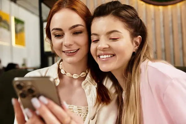 Duas jovens mulheres aproveitam seu tempo em um café chique, sorrindo e olhando para um telefone juntos. — Fotografia de Stock