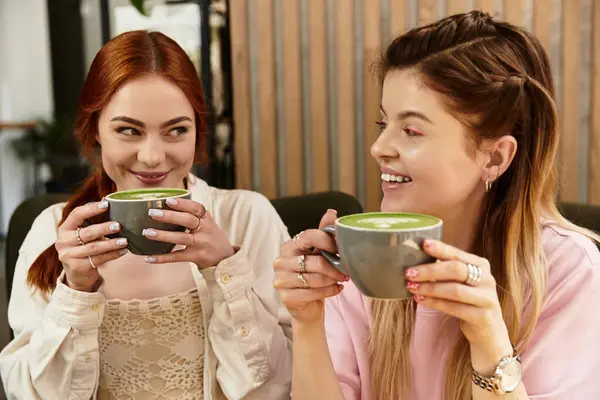 Two young women share laughter and green drinks in a modern cafe setting, embracing friendship. — Stock Photo
