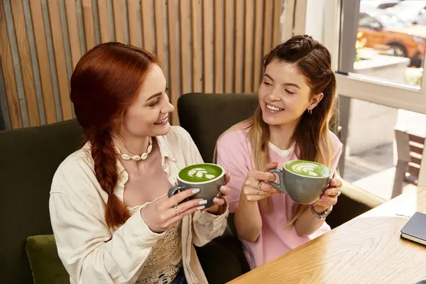 Duas jovens mulheres compartilham um momento alegre enquanto bebem lattes verdes em um café moderno. — Fotografia de Stock