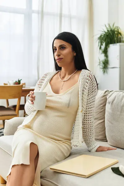 Mulher grávida senta-se confortavelmente no sofá, desfrutando de uma bebida quente em seu sereno espaço de vida. — Fotografia de Stock