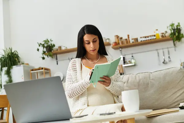 A young, beautiful pregnant woman sits comfortably on a sofa, engrossed in writing in her notebook. — Stock Photo