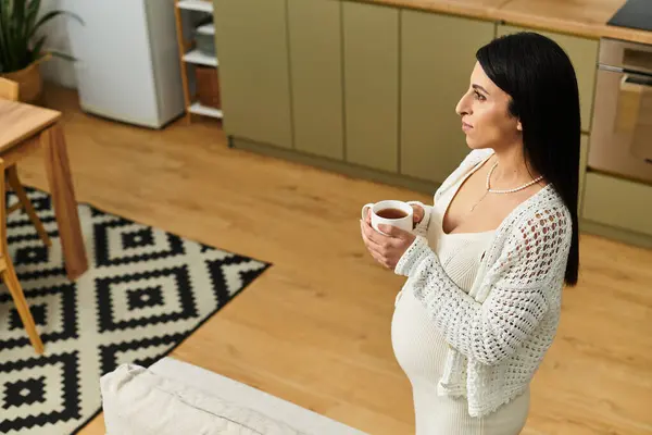 A young pregnant woman stands in her kitchen, savoring tea and lost in thought. — Stock Photo