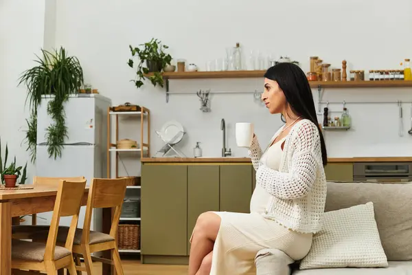 Beautiful pregnant woman relaxes in a cozy kitchen, holding a warm cup of tea and daydreaming. — Stock Photo