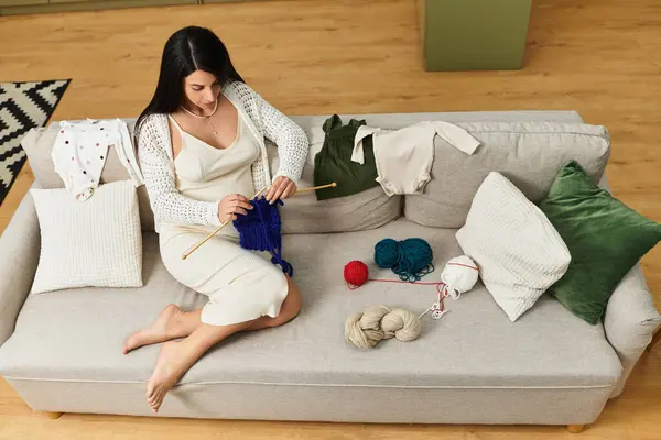 A young expectant mother enjoys knitting on a cozy couch while surrounded by baby clothes and yarn. — Stock Photo