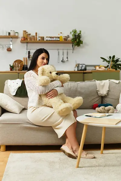 A beautiful young woman in a knitted top holds a teddy bear while sitting on a couch in her home. — Stock Photo