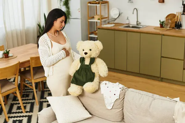 Una hermosa mujer embarazada sostiene con cariño a un osito de peluche en una cocina cálida y acogedora. - foto de stock