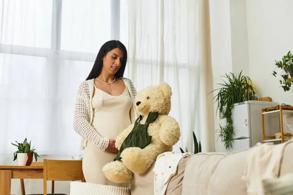 A beautiful pregnant woman gently cradles her baby bump while holding a teddy bear in her home. — Stock Photo
