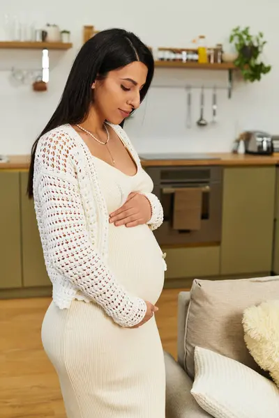 A young beautiful woman gently cradles her baby bump while standing in a warm kitchen, feeling joy. — Stock Photo