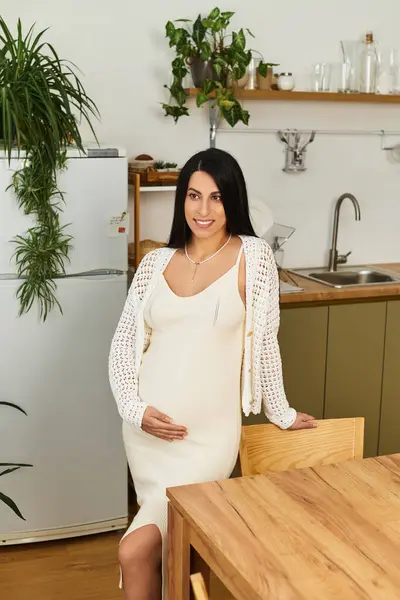 Beautiful pregnant woman sits gracefully in a warm kitchen, showcasing her glowing happiness. — Stock Photo