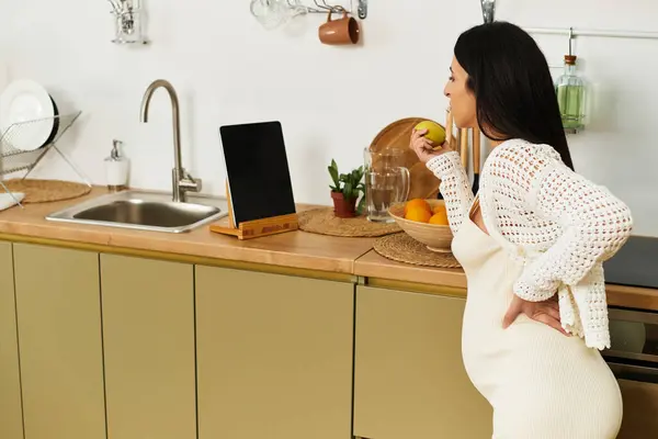 Beautiful young woman with a baby bump stands in a bright kitchen while enjoying an apple — Stock Photo