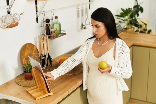 Esperando mãe mantém maçã fresca enquanto navega receita em um ambiente de cozinha elegante. — Fotografia de Stock