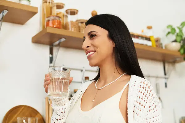 Uma linda mulher grávida sorri enquanto segura um copo de água em sua cozinha quente durante o anoitecer. — Fotografia de Stock