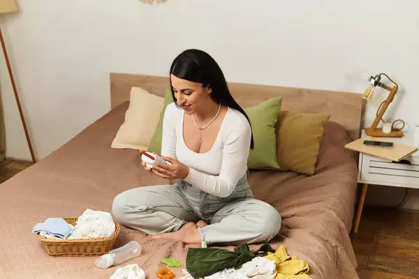 Beautiful young woman lovingly examines baby items while seated on a bed in her warm nursery. — Stock Photo