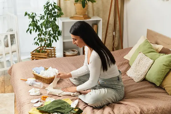 In a serene bedroom, a young woman sits on a bed, organizing baby items with care and joy. — Stock Photo