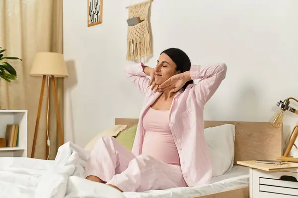 Young woman in cozy pink pajamas stretches joyfully, embracing her pregnancy in a bright bedroom. — Stock Photo