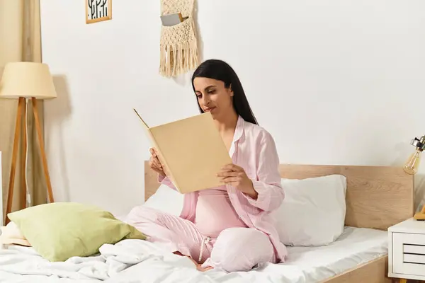 Young beautiful pregnant woman seated cross legged in cozy bedroom, deeply engaged in reading — Stock Photo