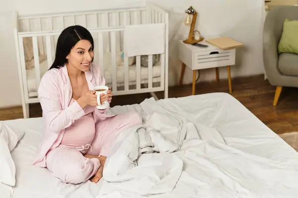 Expecting mother sits comfortably on a bed, sipping a warm drink with a joyful expression. — Stock Photo