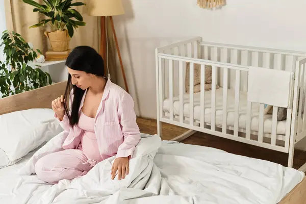 Expecting mother in pink pajamas sits thoughtfully on bed surrounded by soft blankets and a nursery. — Stock Photo