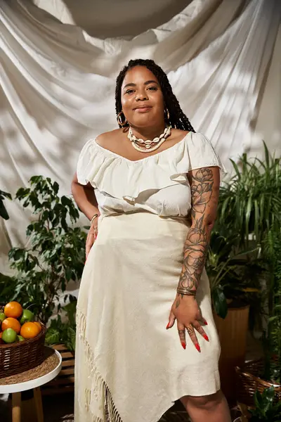 A stylish African American woman in a vibrant summer dress poses gracefully among lush green plants. — Stock Photo