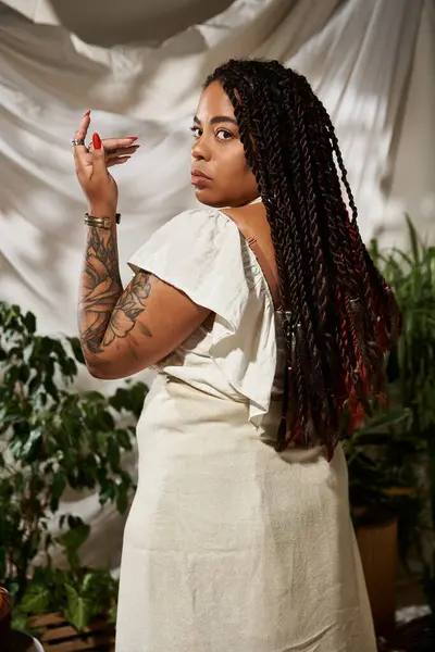 A confident woman with beautiful braids poses elegantly in a summer dress surrounded by greenery. — Stock Photo