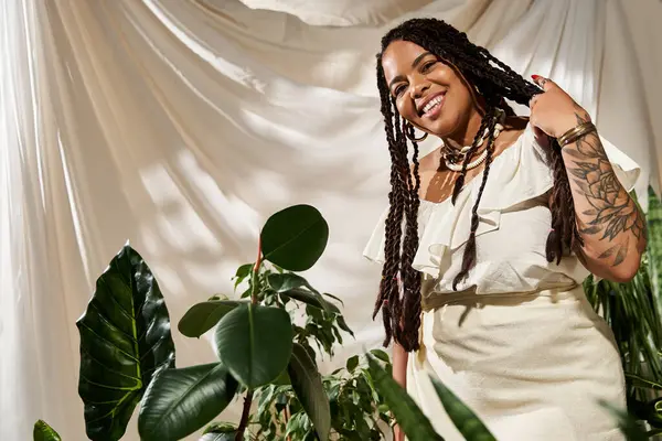 Una mujer alegre con trenzas lleva un vestido de verano y posa en medio de una exuberante vegetación en un estudio luminoso. - foto de stock