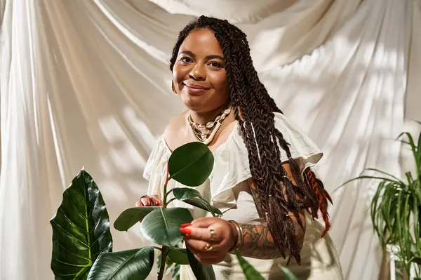 A joyful woman with braids poses in a light summer dress surrounded by vibrant green plants. — Stock Photo