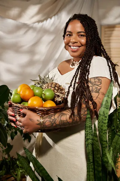 A joyful woman with braids poses with a basket of fresh fruit amidst greenery. — Stock Photo