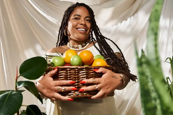 Una donna nera radiosa posa con grazia con un cesto di frutti vivaci in una vegetazione lussureggiante. — Foto stock