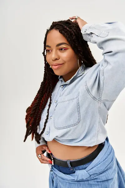 Una donna sicura di sé con le trecce posa in un abito di jeans su uno sfondo grigio, irradiando gioia. — Foto stock